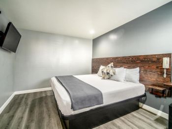 The image shows a modern hotel room with a large bed, gray and white decor, a wall-mounted TV, and a wooden headboard with side shelves.