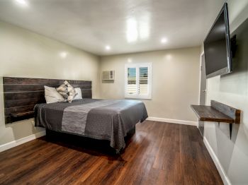 A modern bedroom with wooden flooring, a bed with gray bedding, wall-mounted TV, air conditioner, and a small window, well-lit environment.