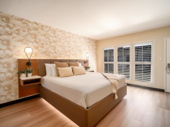 A modern bedroom with a large bed, decorative wall, bedside lamps, plants, and windows with shutters fills the space.