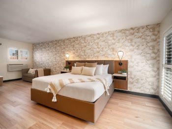 The image shows a modern, well-lit bedroom with a large bed, decorative wall, wooden flooring, bedside tables, a sofa, and a window with blinds.