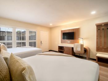 The image shows a hotel room with two beds, a wall-mounted TV, a chair, and a window with blinds. The decor is modern and minimalist.