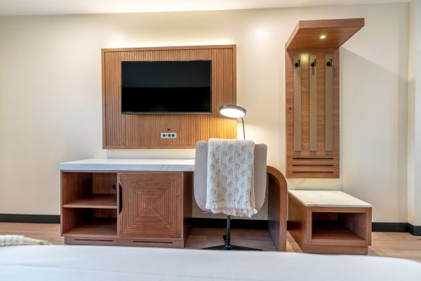 A modern hotel room features a wall-mounted TV, desk with a lamp, chair with a draped towel, and a wooden bench with coat hooks.