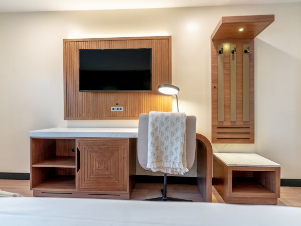 A modern hotel room features a wall-mounted TV, desk with a lamp, chair with a draped towel, and a wooden bench with coat hooks.