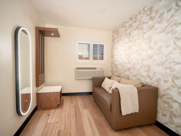 A cozy, small room with a couch, a wall mirror, an air conditioner, and patterned wallpaper. The space appears well-lit and minimally decorated.