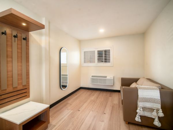 A small room with a brown couch, white blanket, wall mirror, coat rack, bench, wooden floor, window with shutters, and wall-mounted air conditioner.