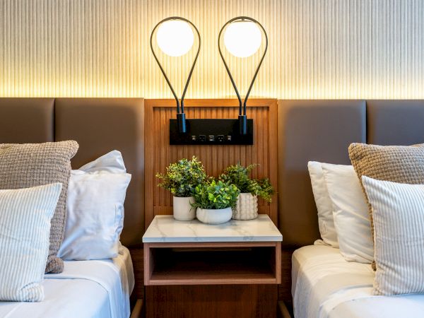 A hotel room features two beds with a shared nightstand holding potted plants. Two modern lamps are mounted above the nightstand, illuminating the area.