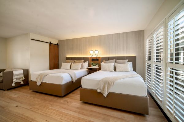 A modern hotel room features two double beds, wooden flooring, a sliding barn door, a small seating area, and large windows with shutter blinds.