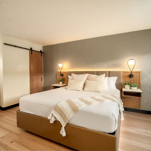 A modern bedroom with a large bed, minimalist decor, wooden flooring, a barn door, and two bedside tables with lamps.