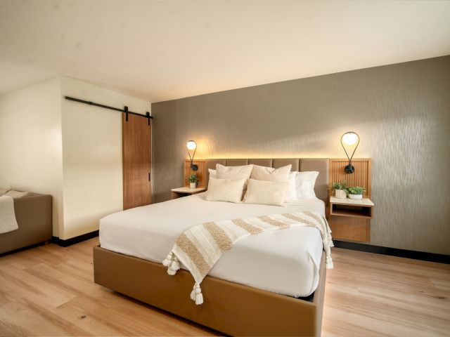A wide-angle view of a hotel room with a king-sized bed, wooden floor, and sliding barn door.