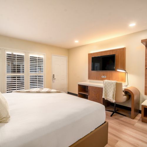 A modern hotel room with a large bed, wall-mounted TV, desk, chair, and ample lighting. White louvered windows and a wooden accent wall are present.