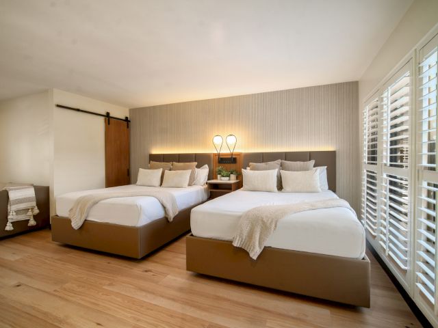 A wide view of a hotel room with two beds, wood floors, and a window with shutters.