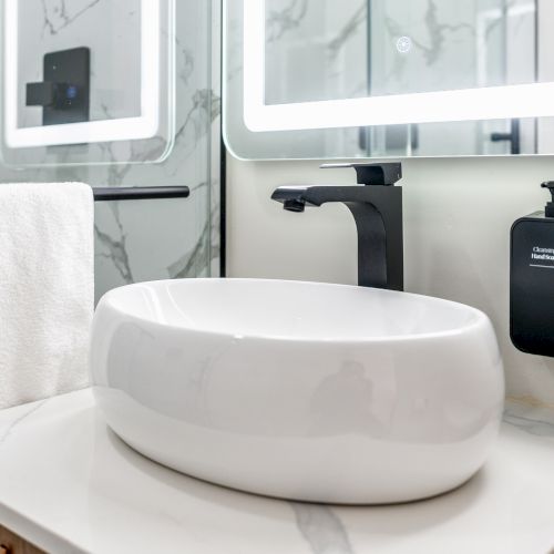 A modern bathroom sink area features a sleek black faucet, towel on a rail, illuminated mirror, and wall-mounted soap dispensers.