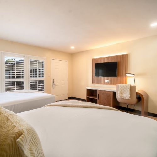 The image shows a modern hotel room with two beds, neutral decor, a wall-mounted TV, a desk, a chair, and natural light from windows with shutters.