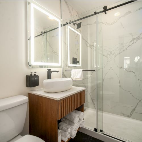 A modern bathroom featuring a toilet, a vanity with a vessel sink, LED-lit mirrors, and a glass-enclosed shower with marble-style walls.