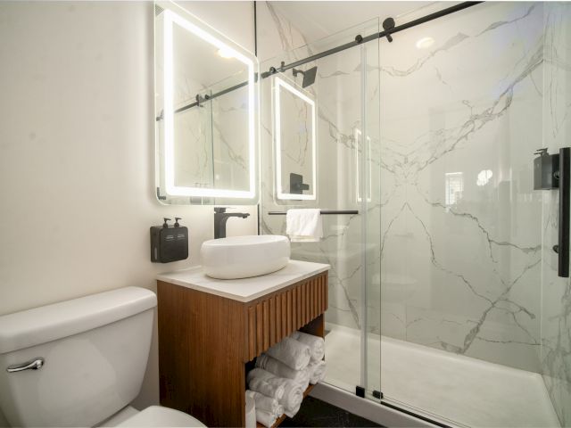 A modern bathroom with a glass-enclosed shower, marble walls, a wooden vanity, and a LED-lit mirror.