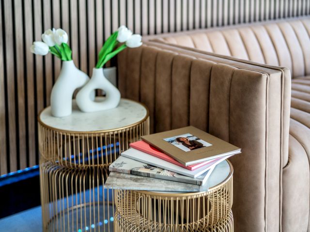 Two small round tables with gold accents and marble tops, one holding two white vases with flowers and the other with a stack of books, in front of a beige couch.