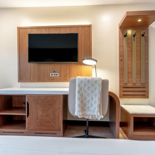 A modern hotel room features a wall-mounted TV, wooden furniture, a desk with a chair and lamp, and a coat rack with a small bench.