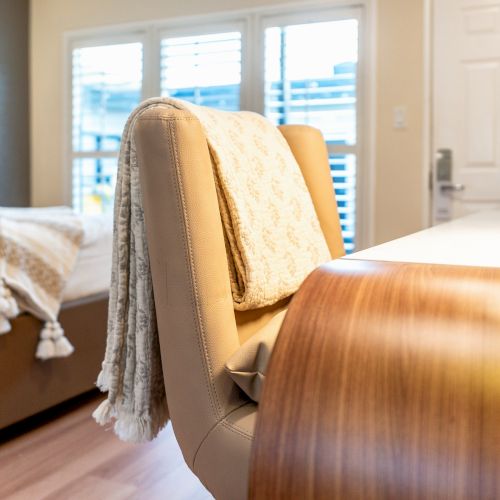 A cozy bedroom with a bed, a chair draped with a blanket, a desk, a lamp, and a door in the background, lit by natural light from windows.