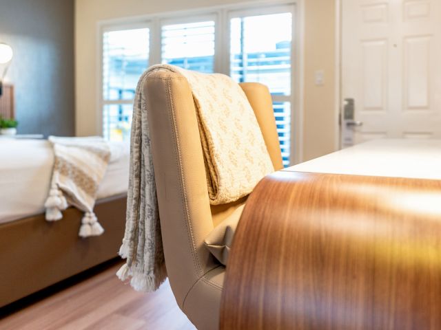 A hotel room with a leather chair, wood table, bed, window, and door.