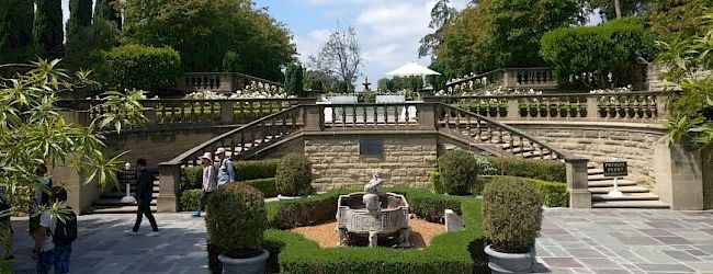 A well-manicured garden with symmetrical stairs, green shrubs, trees, a central sculpture, and people enjoying the area on a sunny day.