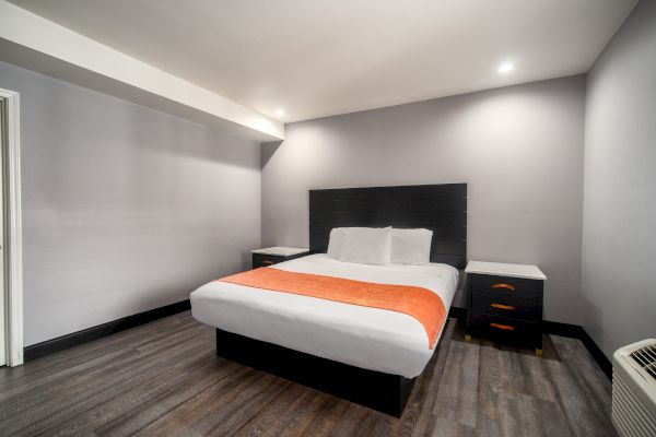 The image shows a modern hotel room with a neatly made double bed, featuring a white and orange color scheme, and simple, stylish furniture.