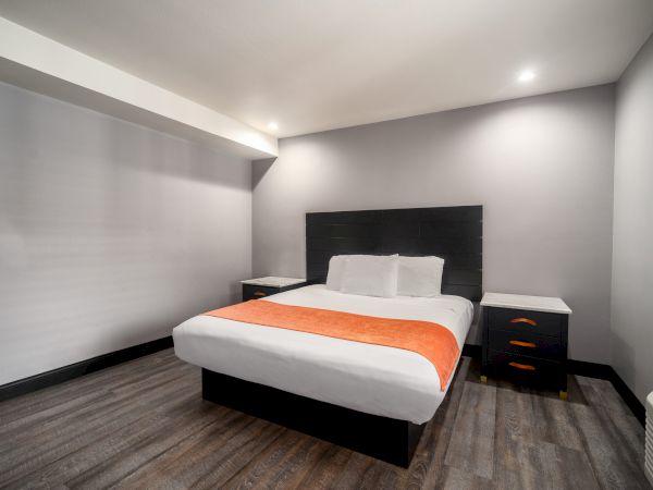 The image shows a modern hotel room with a neatly made double bed, featuring a white and orange color scheme, and simple, stylish furniture.