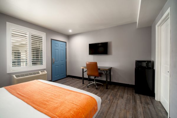 A modern hotel room with a bed, orange blanket, desk, chair, wall-mounted TV, mini-fridge, blue door, and window with shutters.