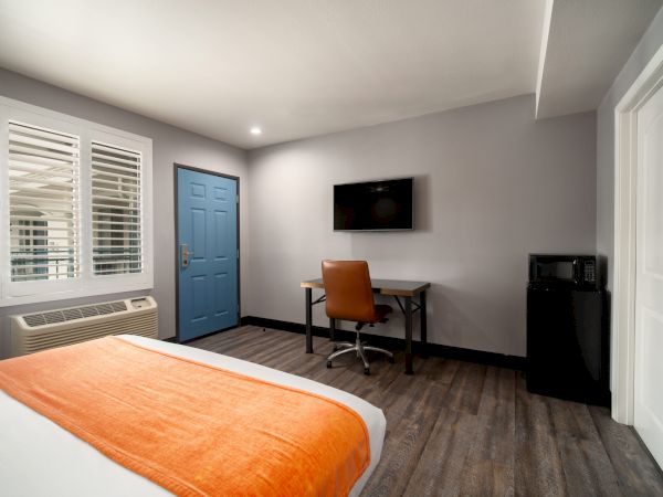 A modern hotel room with a bed, orange blanket, desk, chair, wall-mounted TV, mini-fridge, blue door, and window with shutters.