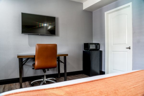 The image shows a room with a wall-mounted TV, a desk with a brown chair, and a small refrigerator with a microwave on top next to a door.