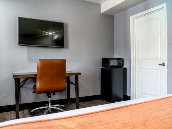 The image shows a room with a wall-mounted TV, a desk with a brown chair, and a small refrigerator with a microwave on top next to a door.