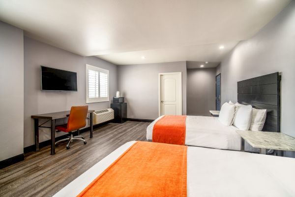 A modern hotel room with two beds, orange blankets, a desk with chair, flatscreen TV, and neutral decor. The room is well-lit and spacious.