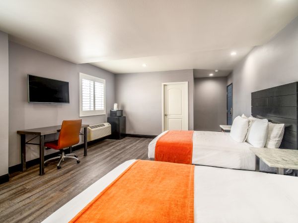 A modern hotel room with two beds, orange blankets, a desk with chair, flatscreen TV, and neutral decor. The room is well-lit and spacious.