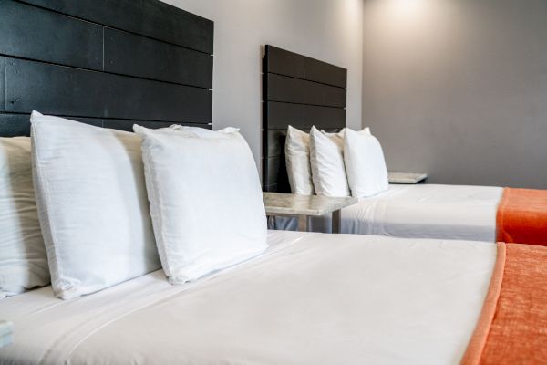 The image shows two neatly made beds with white linens and multiple pillows, against a grey wall with dark headboards, and orange foot runners.