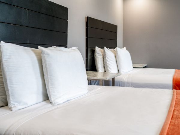 The image shows two neatly made beds with white linens and multiple pillows, against a grey wall with dark headboards, and orange foot runners.
