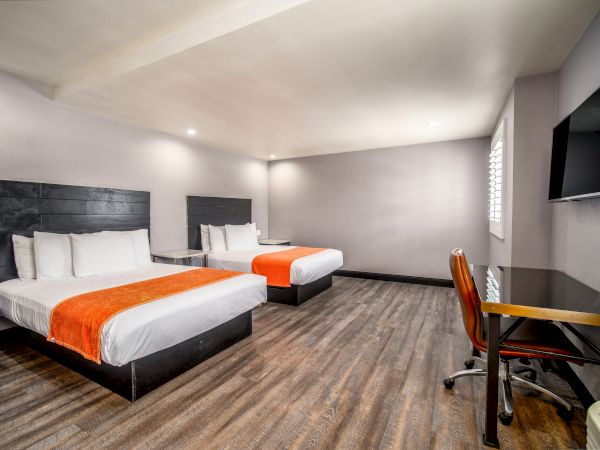 A clean, modern hotel room features two double beds with orange accents, a flat-screen TV, a desk with a chair, and wood flooring, ending the sentence.