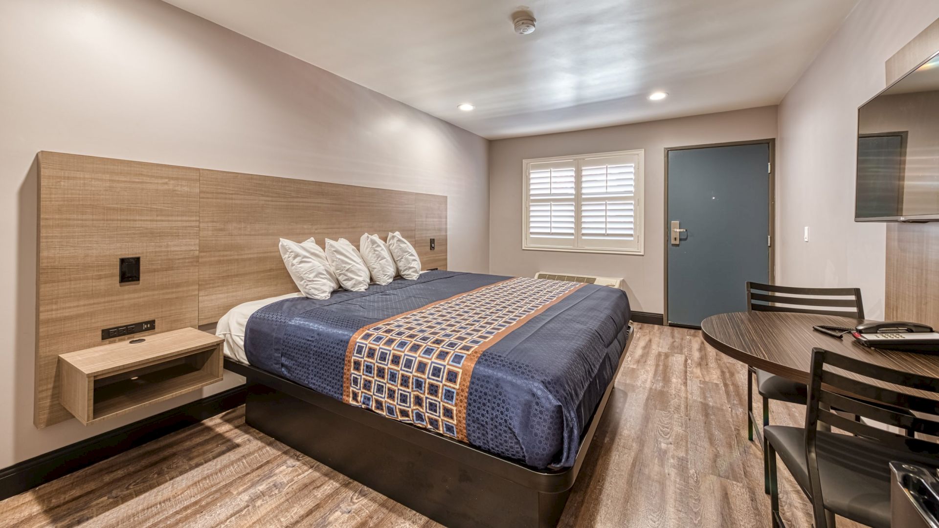 The image shows a modern hotel room with a large bed, wall-mounted nightstand, desk and chair, wooden flooring, and a door with window shutters.