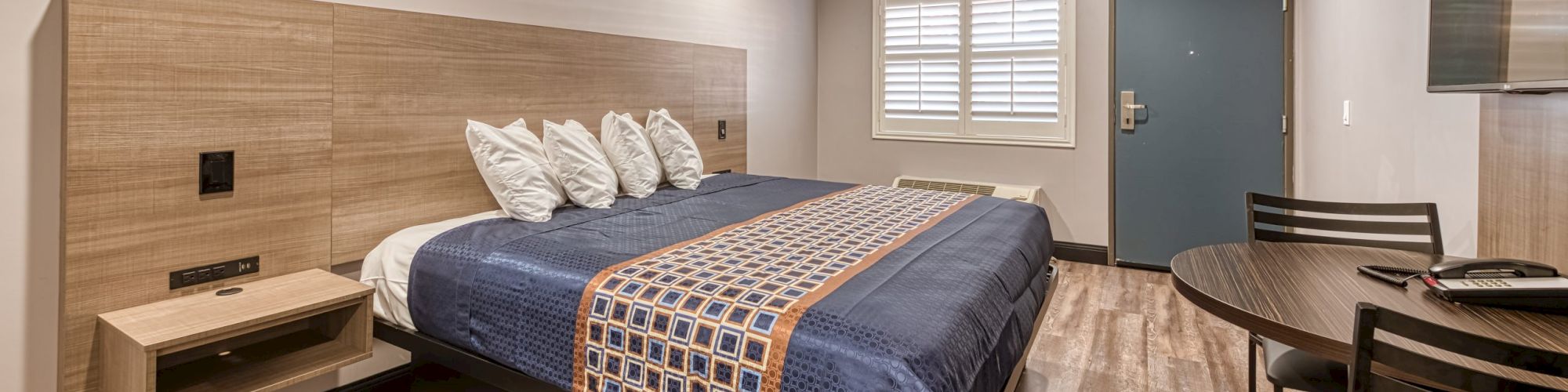 The image shows a modern hotel room with a large bed, wall-mounted nightstand, desk and chair, wooden flooring, and a door with window shutters.