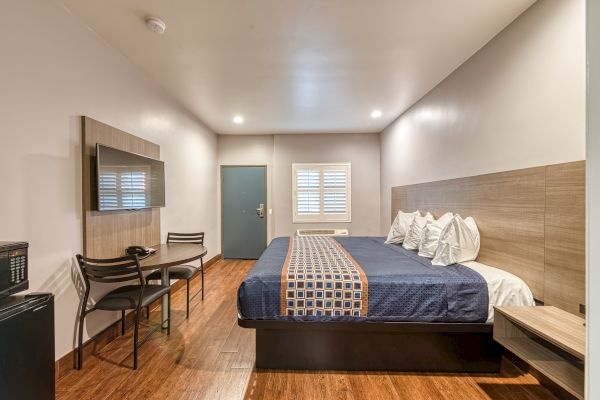 A neatly arranged hotel room with a large bed, table and chairs, wall-mounted TV, window with blinds, and clean, modern decor, viewed from one corner.