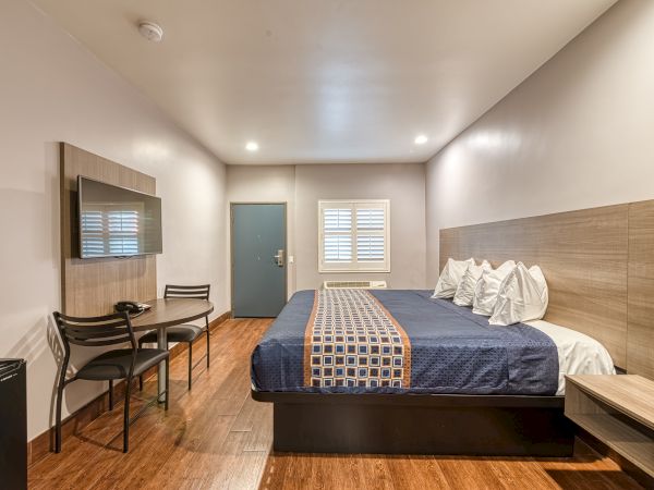 A neatly arranged hotel room with a large bed, table and chairs, wall-mounted TV, window with blinds, and clean, modern decor, viewed from one corner.