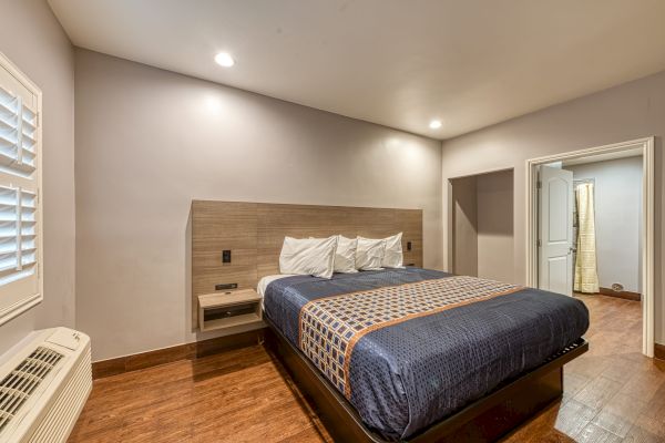 The image depicts a cozy bedroom with a neatly made double bed, wooden floor, nightstands, and bright lighting, also featuring a partially open door.