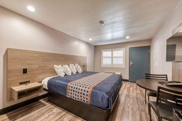 A modern hotel room with a bed, bedside tables, a table with chairs, a wall-mounted TV, and a door with a window. The decor is minimalist.