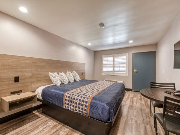 A modern hotel room with a bed, bedside tables, a table with chairs, a wall-mounted TV, and a door with a window. The decor is minimalist.