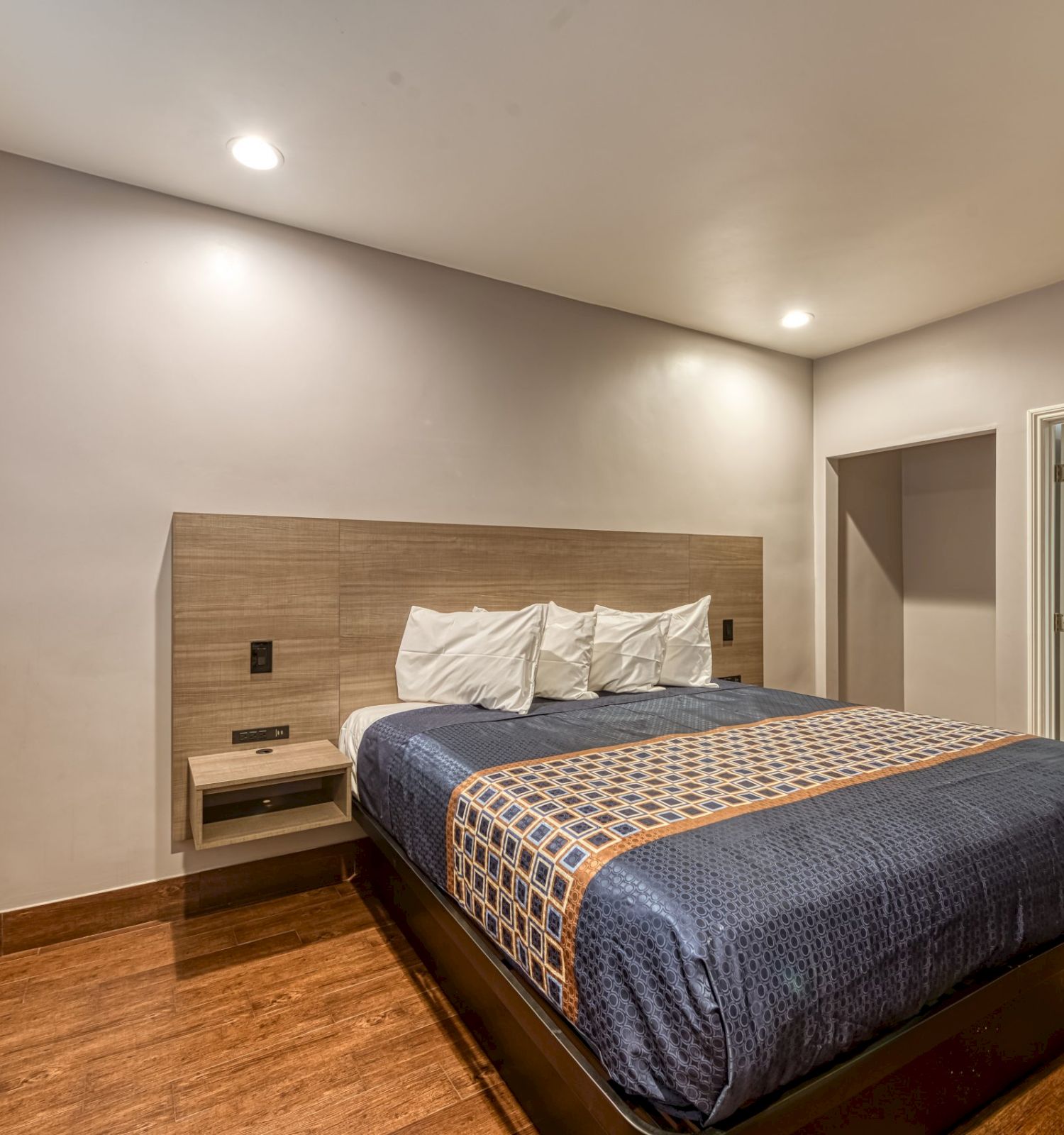 A bedroom with a neatly made bed, wooden floor, bedside table, built-in air conditioner, and an open door to another room.