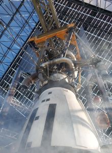 The image shows a rocket engine with piping and structural support inside an industrial or testing facility, with visible steam or smoke.