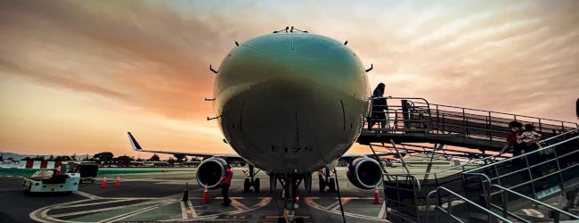 A large aircraft is parked on the tarmac at sunset, with ground crew performing maintenance and a boarding staircase positioned at a door.