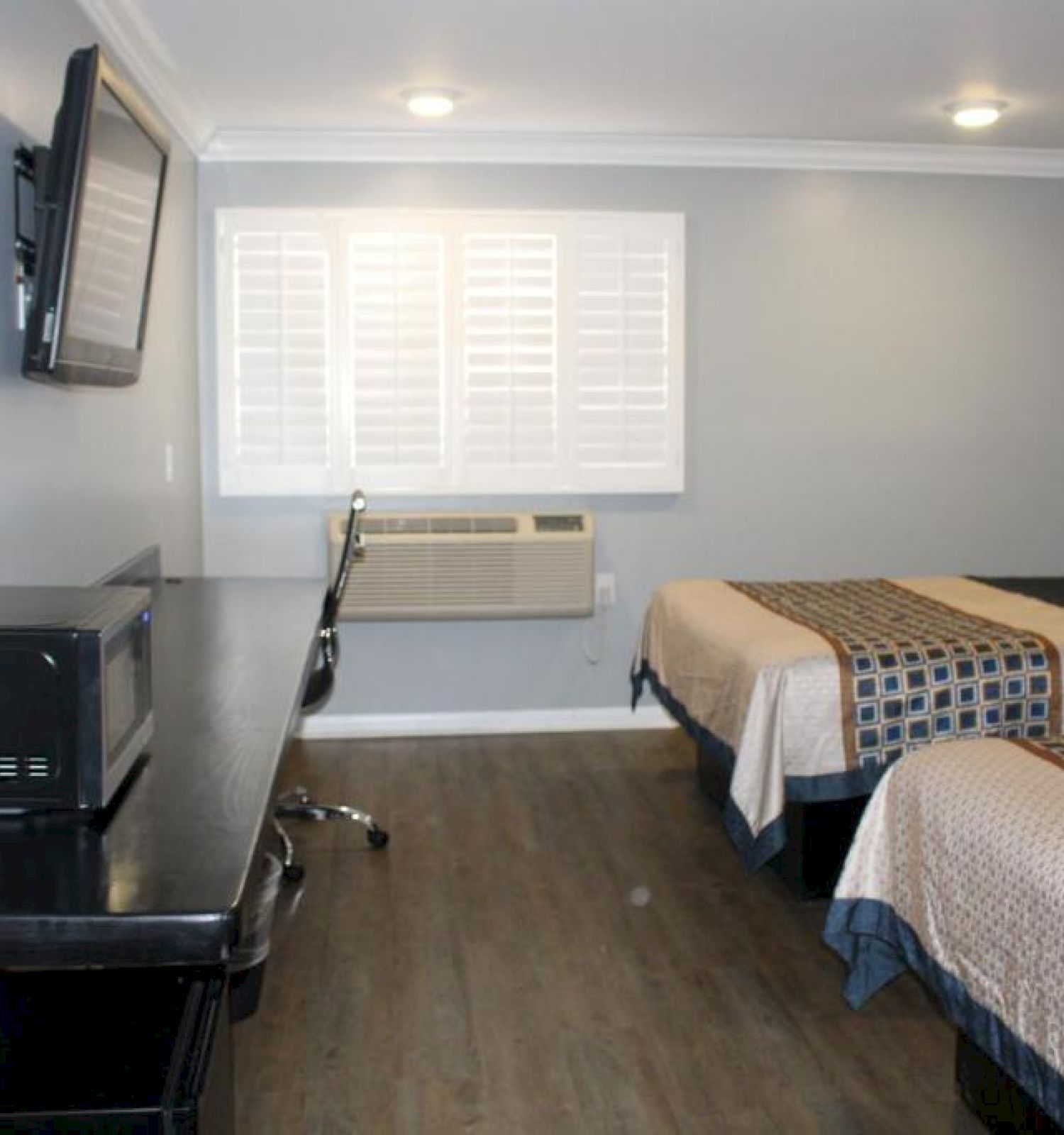 A hotel room with two beds, a wall-mounted TV, a desk with a microwave, air conditioning, and a window with white blinds at the back.