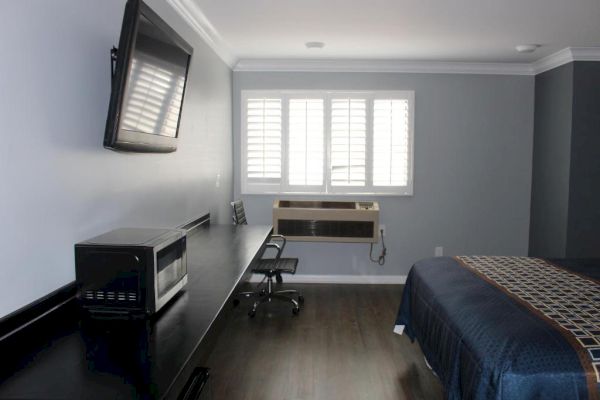 A modern hotel room with a bed, desk, chair, wall-mounted TV, microwave, and window with blinds. The decor is minimalist with neutral tones.