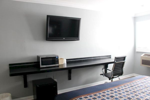 The image shows a minimalist room with a wall-mounted TV, a black desk holding a microwave and a tissue box, a rolling chair, and a window.