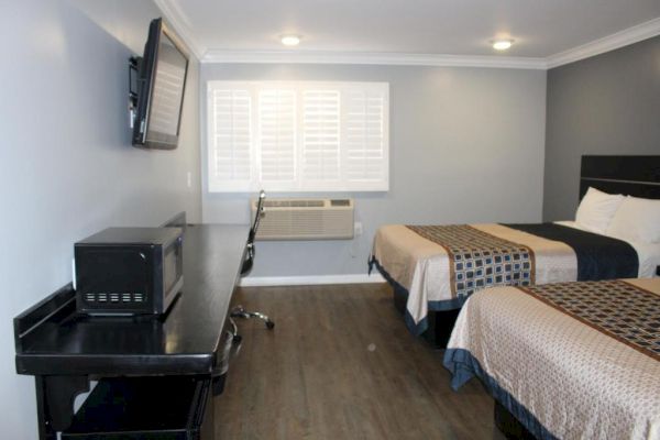 The image shows a hotel room with two beds, a flat-screen TV, a desk with a microwave on it, and a window with shutters.