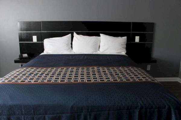 A neatly made bed with three white pillows, a blue blanket, and a patterned runner, set against a dark headboard and a gray wall.